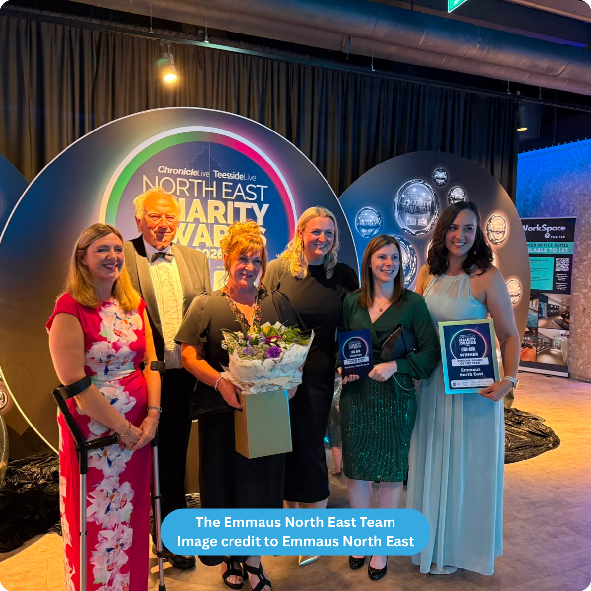 Group of 1 man and 5 women posing for a photo, holding a bunch of flowers and 2 award frames, against an oval shaped background stating North East Charity Awards