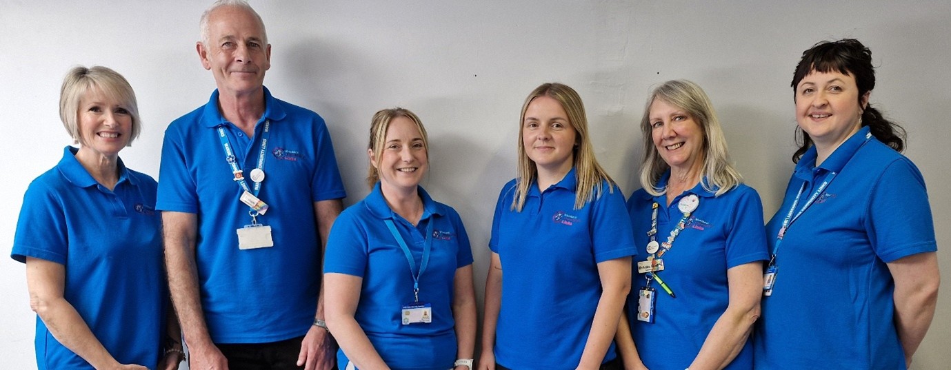 Six members of the team smiling at camera and wearing thier blue NHS branded uniform tops and colourful lanyards with lots of badges hooked on the cords.